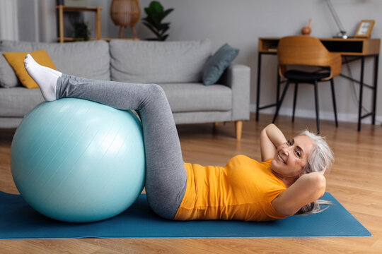 Sporty senior lady exercising her abs muscles with fitball on yoga mat at home, looking and smiling at camera