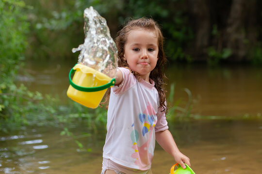 Little girl throwing water with a bucket into the river. Girl with swimsuit and t-shirt. Children's photo in summer.