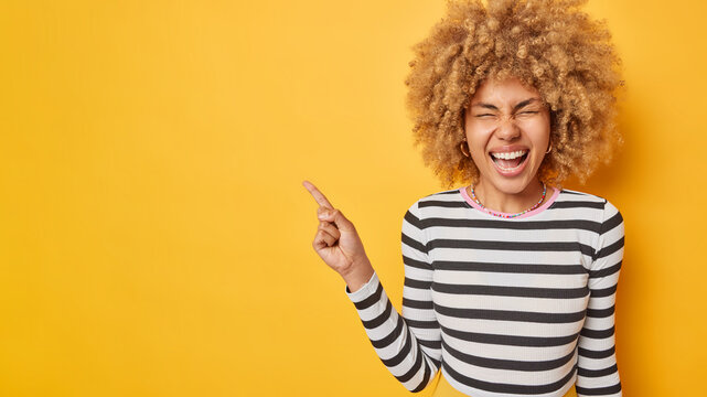 Overjoyed happy young woman with curly blonde hair points away on blank space advertises something laughs gladfully wears casual striped jumper isolated over vivid yellow background. Look there