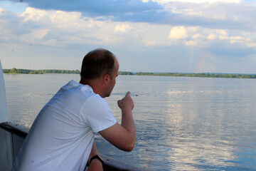 a man with a cigarette in his hand stands on the deck of a ship and points to a rainbow in the sky