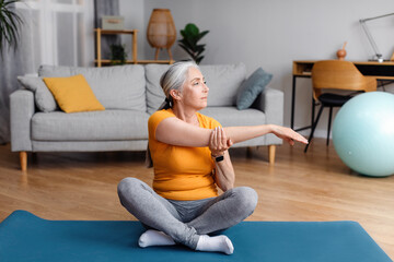Active senior woman stretching her arm on mat at home, doing flexibility exercises, practicing yoga or pilates