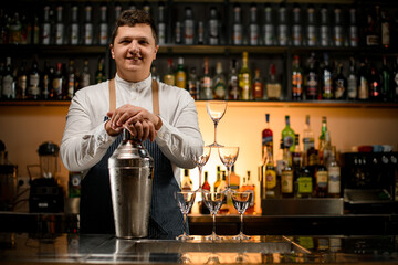 smiling man bartender holds large shaker and pyramid of empty glasses stands nearby on bar