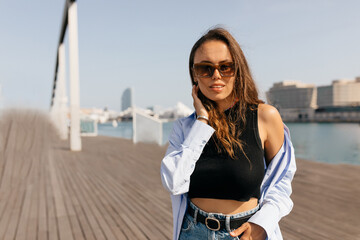 Obraz premium Good-looking stylish girl with long wavy hairstyle wearing black top and blue shirt in sunglasses posing in sunlight on the pier with background of lake