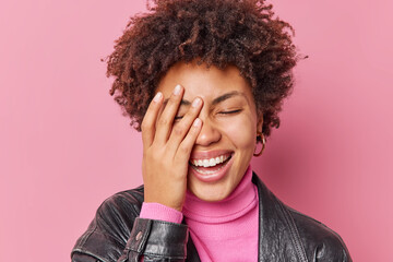 Young curly haired woman makes face palm laughs joyfully expresses positive emotions hears funny...