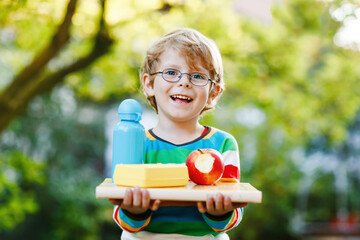 Happy little preschool boy with books, apple and drink bottle on his first day to elementary school or nursery. Smiling child, student with glasses, outdoors. Back to school education concept.