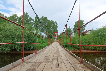 Suspension wooden pedestrian bridge across the river with overgrown banks. Russia, Arkhangelsk, Solombalka River