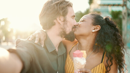 Closeup of smiling interracial couple eating ice cream and taking a selfie on urban city background. Close-up of a man and woman tasting ice cream and video chatting using a mobile phone. Backlight