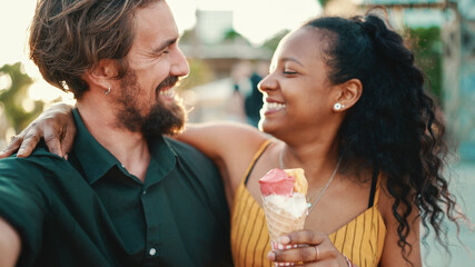 Closeup of smiling interracial couple eating ice cream and taking a selfie on urban city background. Close-up of a man and woman tasting ice cream and video chatting using a mobile phone. Backlight