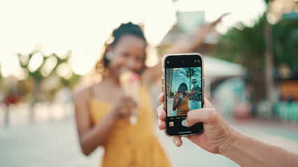 Close-up of a man taking a picture of a woman on a mobile phone. Closeup, smiling woman eating ice cream and filming it with smartphone on urban city background. Backlight