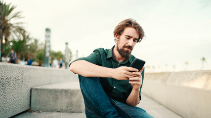 Fototapeta premium Close-up portrait of a man with a beard recording a voice message on the embankment background. Closeup of a young hipster male using a mobile phone
