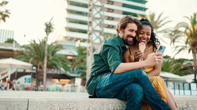 portrait of happy man and smiling woman watching video on smartphone. Close-up of joyful young interracial couple browsing photos on mobile phone on urban city background