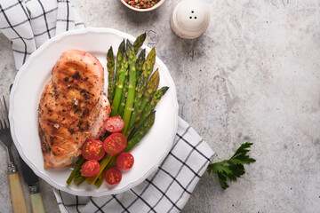 Chicken breast grilled with vegetables. Grilled chicken steak, asparagus and cherry tomatoes in black plate on light gray concrete table background. Healthy diet lunch. Barbecue steak fried. Top view.