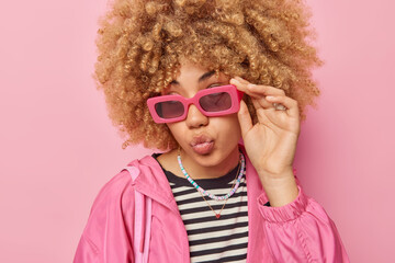 Romantic beautiful woman with curly hair keeps lips folded keeps hand on sunglasses wears casual...