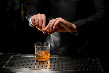 Hands of barman accurate splashing orange peel juice into cocktail in dark bar
