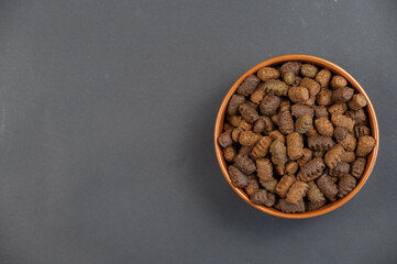 Dry dog food in a brown clay bowl against a gray background. The pellets are oval shaped with jagged edges. Food for senior pets. Useful pet food concept. Top view. Copy space