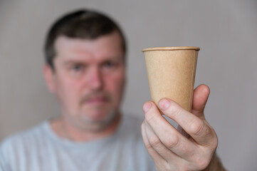 A middle-aged man holds a brown glass in front of his face. A disposable paper drink glass in his outstretched hand. Person with brown short hair. Front Focus. Inside the room.