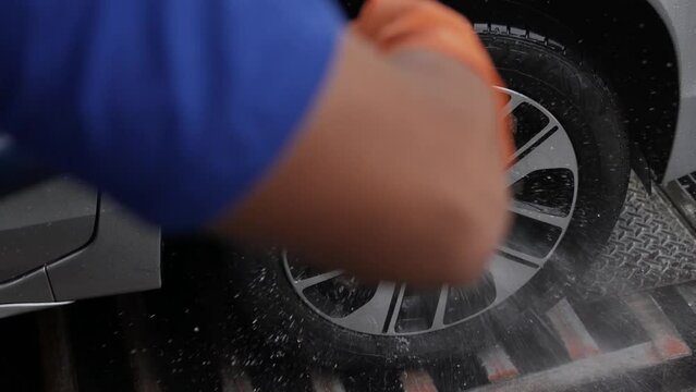 Close up of car wash worker wearing protective gloves and washing car's weel with using hose. Cleaning services.