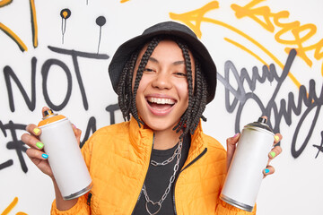 Positive hipster girl with braided hair holds two aerosol sprays draws graffiti on generic wall...