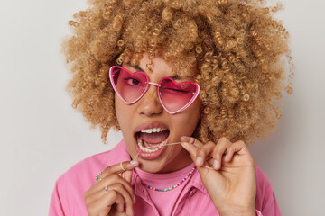 Close up shot of curly haired woman winks eye wears pink sunglasses and jacket uses dental floss...
