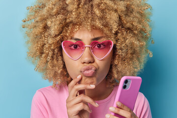 Headshot of serious attentive woman with curly hair keeps lips folded looks directly at camera uses smartphone thinks on downloading new app wears sunglasses and casual t shirt isolated on blue