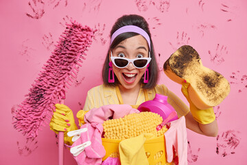 Positive surprised Asian maid wears sunglasses headband rubber gloves performs domestic duties uses...