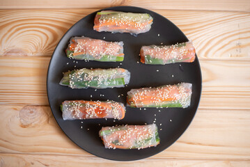Raw spring rolls served on a black ceramic plate on wooden background, flat lay food photo