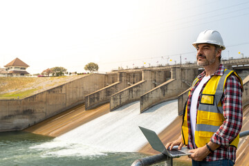 A dam engineering doing his checking routine. He is wearing a white hard hat and yellow transparent vest. He is standing by the rail by the dam.
