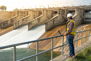 A dam engineering doing his checking routine. He is wearing a white hard hat and yellow transparent vest. He is standing by the rail by the dam. - obrazy, fototapety, plakaty