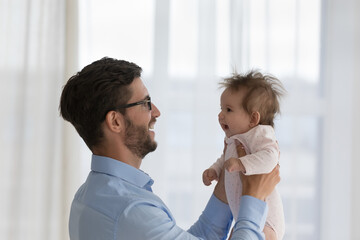 Young loving daddy in glasses standing indoor holds lifts on hands his cute baby, spend priceless time together with infant girl, side profile view. Happy Father Day, fatherhood, family ties concept