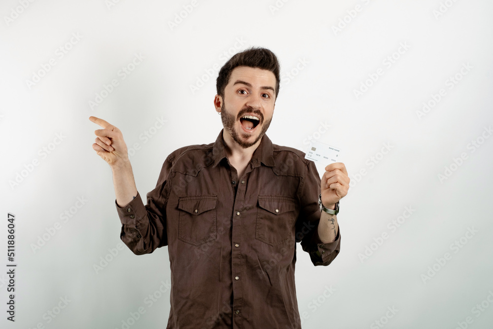 Wall mural Portrait of young caucasian man wearing shirt posing isolated over white background holding credit bank card and pointing index finger aside. Shopping and finance concept.