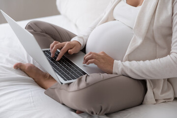 Cropped close up shot gravid woman sit on bed with laptop, future mom buy goods on-line, purchase clothes for unborn baby on internet, e-shopping, client of distance e-services for pregnant concept