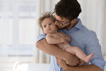 Loving dad kisses on cheek his adorable baby, standing at home holding on arms infant in diaper spend time together, feeling love, express caress and care. Happy fatherhood, babyhood, cherish concept