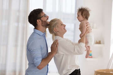 Overjoyed young couple enjoy playtime, spend leisure standing in modern nursery play with their baby cute adorable infant in diaper. Family bond, unconditional love, happy parenthood, babyhood concept