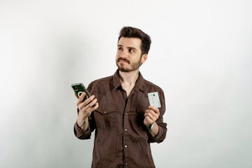 Caucasian young man wearing shirt posing isolated over white background holding smartphone and credit card with anxious expression while looking aside.