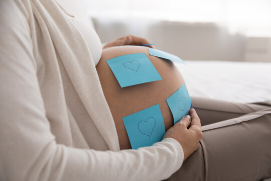 Close up cropped shot unknown pregnant woman touch her big bare belly with attached blue post-it sticky notes with drawn hearts symbols. Happy motherhood, its a boy, love, maternity, pregnancy concept