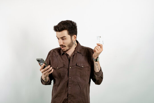 Caucasian man wearing brown shirt posing isolated over white background holding smartphone and credit card shocked with surprise and amazed expression, fear and excited face.