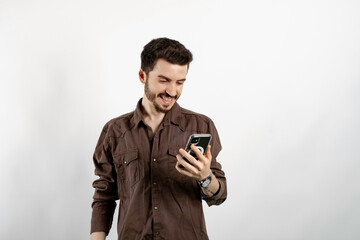 Cheerful caucasian man wearing brown shirt posing isolated over white background looking at screen of smart phone, browsing web pages and smiling while chatting with friends.
