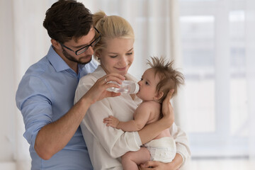 Baby in diaper on parents arms drinks fresh clear water from bottle. Parents keep care of their...