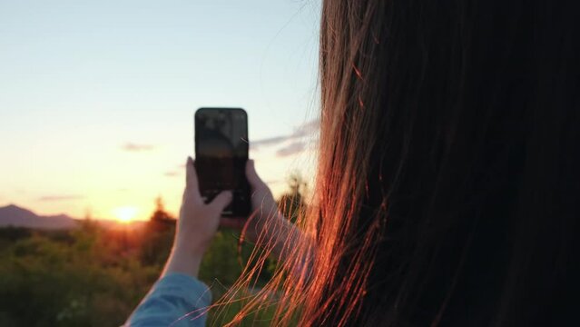 Selective focus of young female holding mobile phone and records a video or photo during epic sunset in amazing beautiful mountains. Capturing sundown on cell phone. Adventure and weekend concept