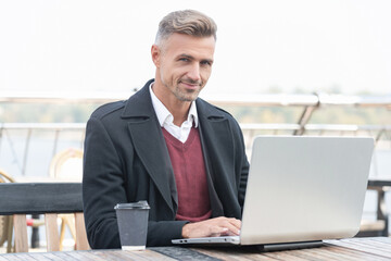 Happy man employee working online in laptop drinking coffee in outdoor cafe