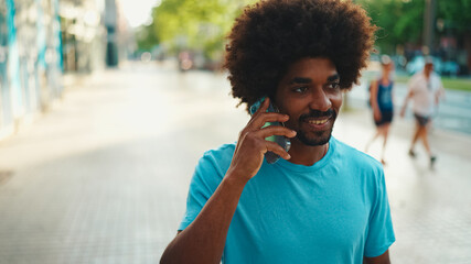 Closeup of smiling young African American man in light blue t-shirt walking down the street and talking on mobile phone