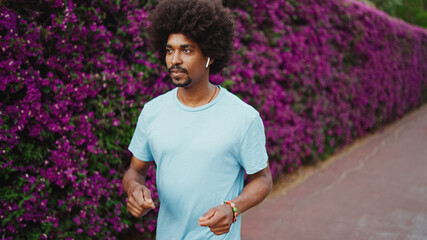 Fototapeta premium Closeup cheerful young African American man listening to music in wireless headphones and dancing on background wall covered with purple flowers. Lifestyle concept.
