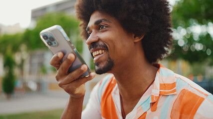 Fototapeta na wymiar Close-up porttrait of young African American man wearing shirt sitting on park bench using his smartphone. Smiling man sends voice message on mobile phone.