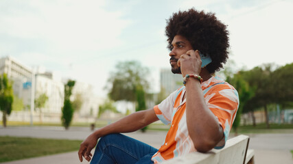Close-up of young African American man wearing shirt sitting on park bench using his smartphone. Smiling man talking on mobile phone.