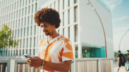 Closeup young African American man wearing shirt writes in social networks on mobile phone on urban street background. Camera moving forwards approaching to the person.