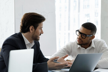 Multi racial businessmen talking during formal meeting in modern office, sit at desk with laptops lead conversation, share solutions looking serious, solve business. Cooperation, negotiations concept