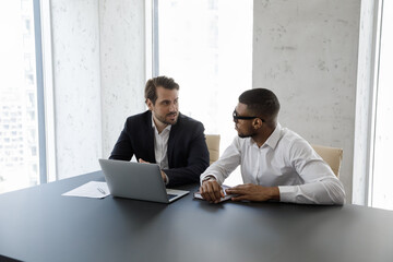 Multi ethnic business partners sit at desk with laptop work on collaborative task or project in modern office, share ideas, express opinion, make solution. Sales manager and client negotiation concept