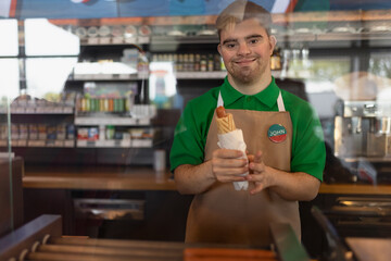 Happy waiter with Down syndrome standing by counter and giving sandwich to a costumer in cafe at gas station.