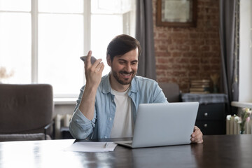 Young businessman sit at desk with laptop holds cellphone listen audio message smile get good news, make call on speakerphone enjoy modern tech for comfort life. Business, workflow, innovation concept