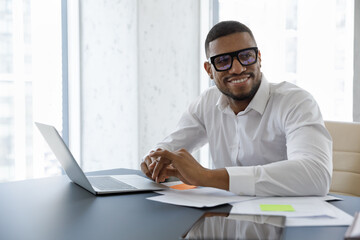 Handsome African businessman sit at desk with laptop smile staring aside in skyscraper office...
